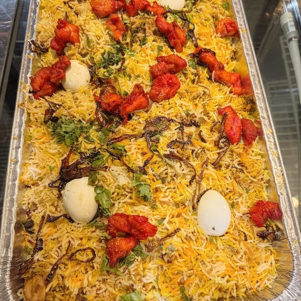 A large tray of Desi Daawat's chicken biryani with red chicken pieces, eggs, and herbs.
